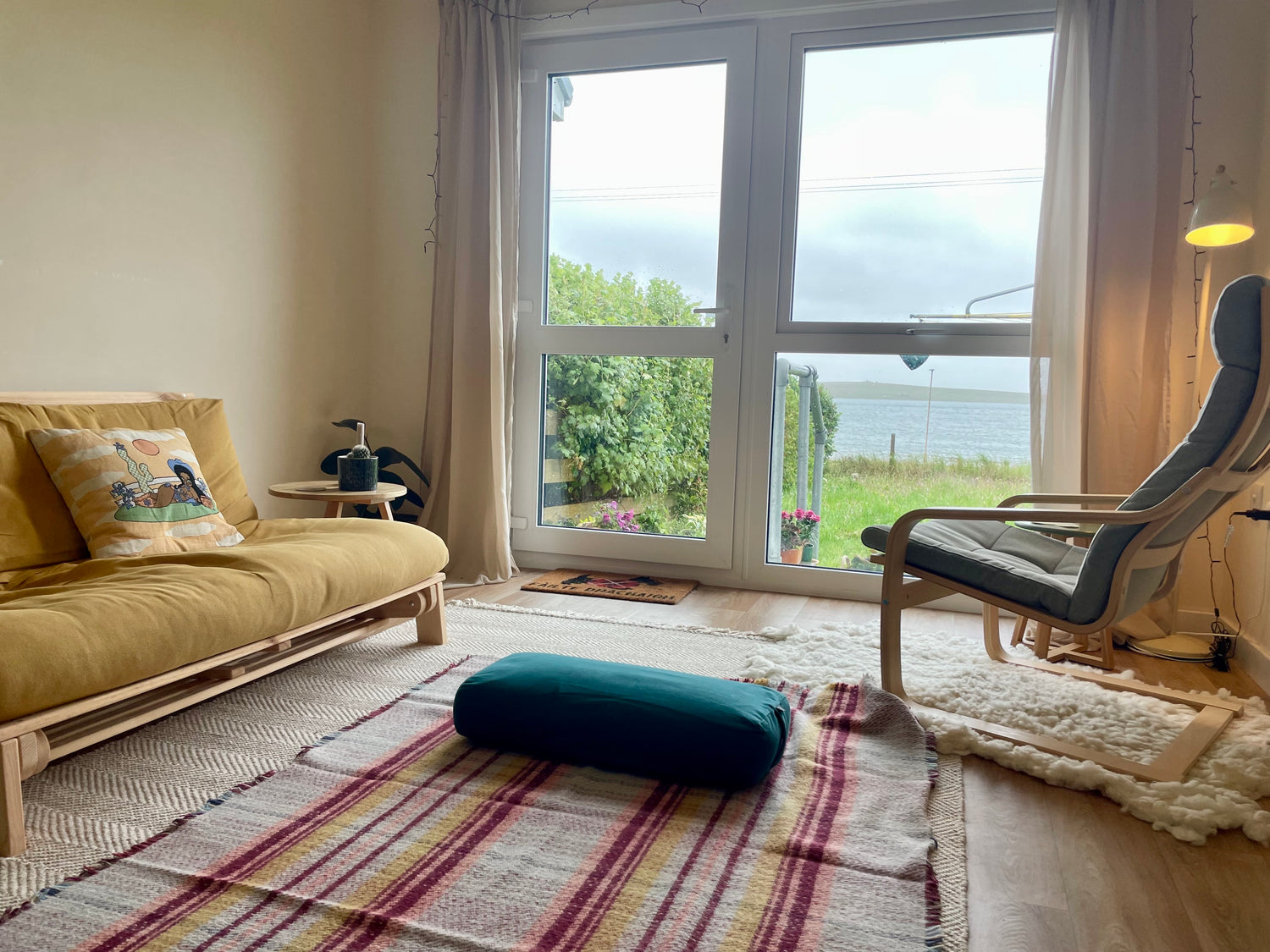 Living room with a yellow sofa, blue bolster, yoga mat and a large window overlooking a garden and the sea.